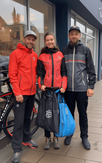 A group of three friends, consisting of two men and one woman, standing outside a local bike shop. Each of them is holding a branded shopping bag from a different cycling store: one from Decathlon, another from Bikester, and the third from Sklep-Rowerowy.Pl. They are engaged in a lively conversation, comparing their recent purchases and sharing their cycling experiences.
